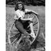 Young woman sitting on a wagon wheel and smiling Poster Print (18 x 24)